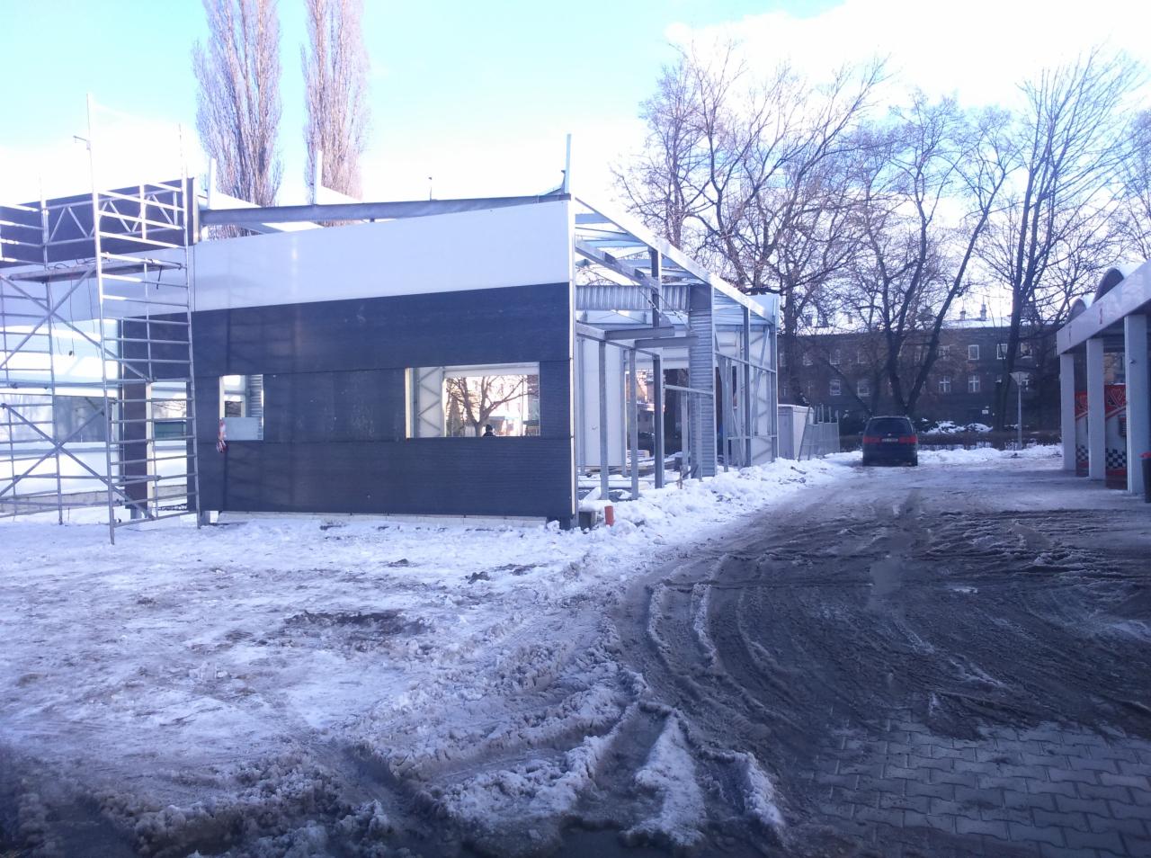 Budowa hali z płyt warstwowych w zimowych warunkach, widoczne metalowe elementy konstrukcyjne i częściowo zamontowane płyty w kolorach białym i grafitowym.