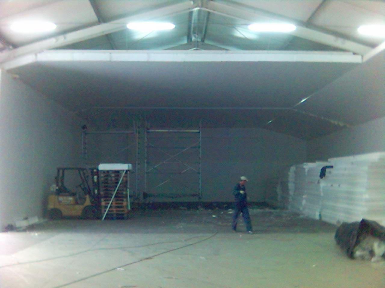 Wnętrze hali z płyt warstwowych w trakcie prac, widoczny wózek widłowy z paletami, pracownik w oddali i składowane płyty.