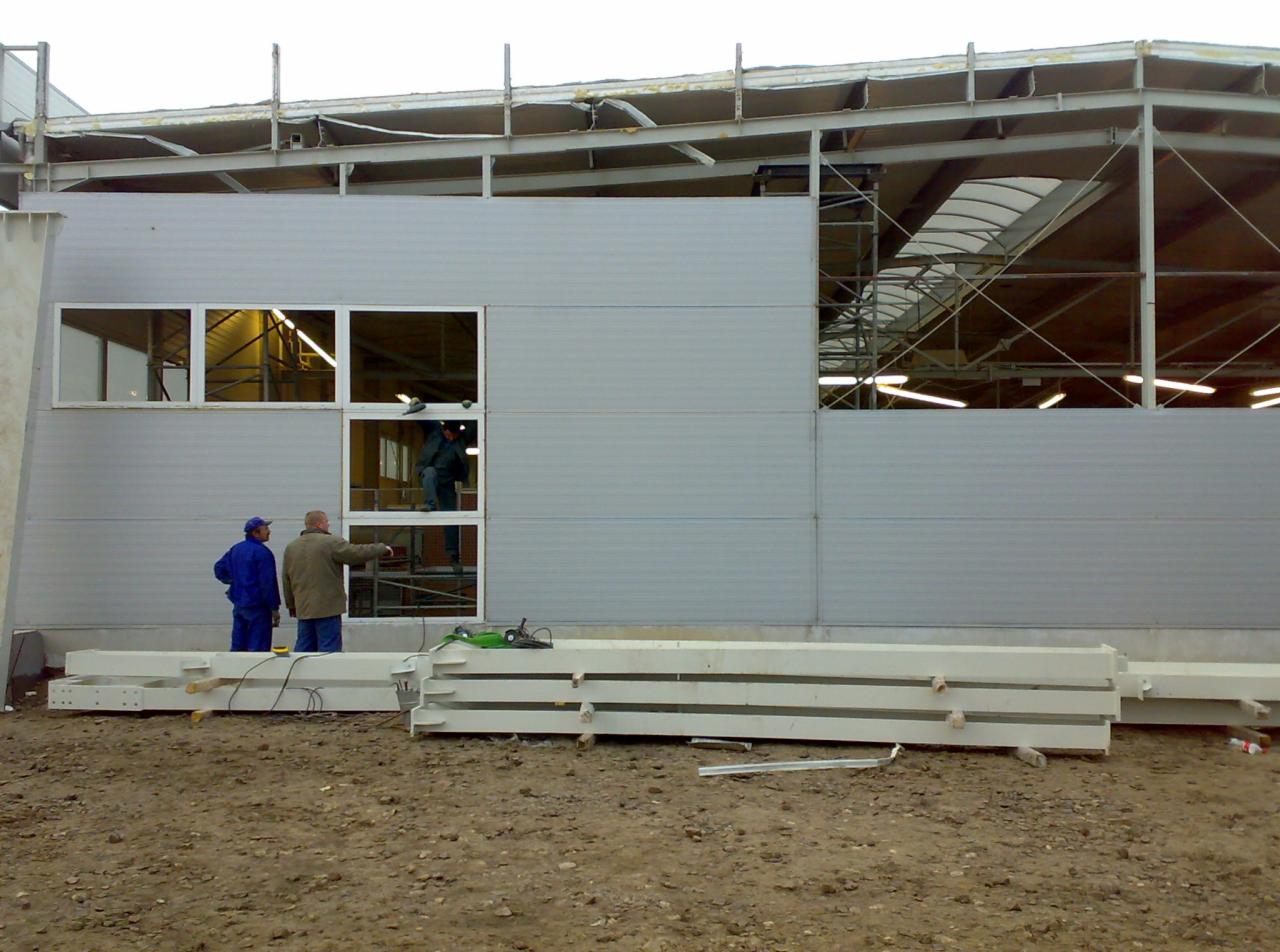 Montaż ściany z płyt warstwowych na konstrukcji stalowej hali. Dwóch mężczyzn na zewnątrz, jeden wewnątrz na rusztowaniu. Na pierwszym planie leżą elementy konstrukcji.