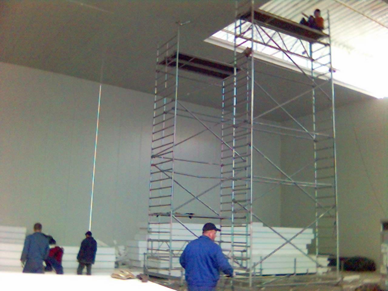 Montaż paneli warstwowych w hali produkcyjnej, widoczne rusztowanie i pracownicy w strojach roboczych.