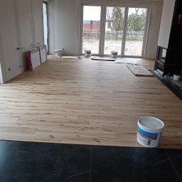 EMS SYSTEM - Wnętrze w trakcie remontu: jasne panele podłogowe ułożone na fragmencie czarnej, marmurowej posadzki, widoczne puszki z farbą i narzędzia, surowe ściany przygotowane do malowania, duże okna...