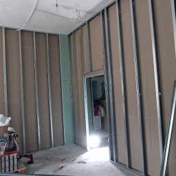 EMS SYSTEM - Wnętrze w trakcie montażu ścian działowych z płyt kartonowo-gipsowych na metalowym stelażu, widoczne przewody wentylacyjne pod sufitem, materiały budowlane i narzędzia na podłodze, oraz fragment...