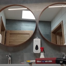EMS SYSTEM - Łazienka z dwoma okrągłymi lustrami z drewnianą ramą nad umywalką, na której leży pistolet do silikonu i inne narzędzia, widoczny dozownik mydła i ręczników papierowych, szare płytki na ścianach.