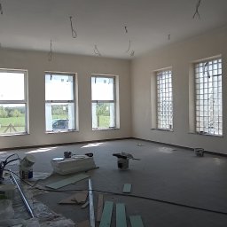 EMS SYSTEM - Wnętrze dużego pomieszczenia w trakcie malowania, widoczne zabezpieczone okna, ślady farby na ścianach, drabina, oraz narzędzia malarskie i płyty izolacyjne na podłodze.