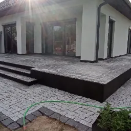 Nowoczesny taras z ciemnej, wzorzystej płyty, schody z tego samego materiału, obrzeża z kostki granitowej, widoczny fragment domu z panelami słonecznymi, trawnik i ogrodzenie.