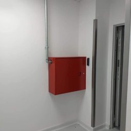EMS SYSTEM - Czerwona szafka hydrantowa zamontowana na białej ścianie w pomieszczeniu z szarą podłogą i widoczną rurą instalacji przeciwpożarowej, z fragmentem ościeżnicy drzwi po prawej stronie.