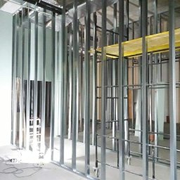EMS SYSTEM - Montaż metalowej konstrukcji szkieletowej ścian działowych z płyt karton gips, widoczne profile pionowe i poziome, fragment rusztowania z żółtą platformą roboczą, instalacje pod sufitem.