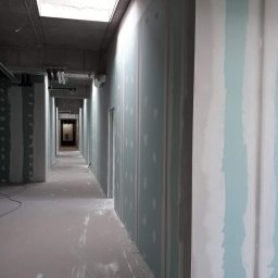 EMS SYSTEM - Długi korytarz w trakcie wykańczania z płyt kartonowo-gipsowych, widoczne łączenia szpachlowane, surowy betonowy sufit z instalacjami, świetlik w suficie, podłoga z resztkami gipsu.