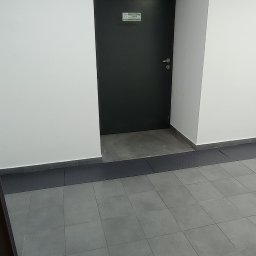 EMS SYSTEM - Szare płytki podłogowe w korytarzu biurowym, widoczna czarna ściana z drzwiami i tabliczką informacyjną.