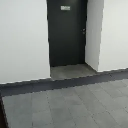 Szare płytki podłogowe w korytarzu biurowym, widoczna czarna ściana z drzwiami i tabliczką informacyjną.