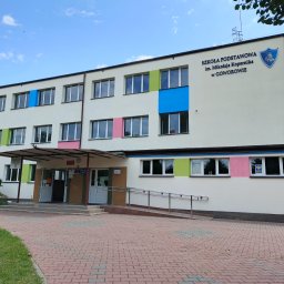 EMS SYSTEM - Fasada budynku szkoły podstawowej z kolorowymi akcentami wokół okien: niebieski, zielony, różowy. Wejście z podjazdem dla wózków. Czerwona kostka brukowa.