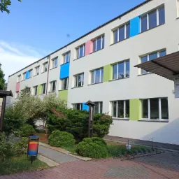 Budynek szkoły po termomodernizacji z kolorowymi obramowaniami okien: niebieskie, zielone, różowe. Widoczny fragment chodnika i zieleń. Tablica informacyjna.