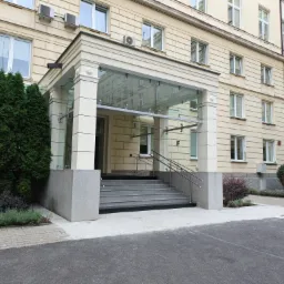 Wejście do budynku z przeszklonym zadaszeniem i granitowymi schodami. Elewacja w odcieniach beżu, widoczne okna, klimatyzatory. Obok wejścia zieleń.