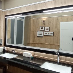 EMS SYSTEM - Umywalnia publiczna z dwoma umywalkami wpuszczonymi w czarny blat, duże lustro w czarnej ramie z oświetleniem LED, dozowniki na mydło i ręczniki, ściany wykończone płytkami imitującymi drewno...