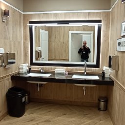 EMS SYSTEM - Wykończona łazienka publiczna z dwoma umywalkami na czarnym blacie, lustrem z podświetleniem LED, drewnopodobnymi płytkami na ścianach i podłodze, oraz elementami dekoracyjnymi jak zdjęcia...