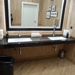 EMS SYSTEM - Nowoczesna łazienka z dwoma umywalkami nablatowymi na czarnym blacie, duże podświetlane lustro z czarną ramą, ściany i podłoga wykończone panelami imitującymi jasne drewno ułożone w jodełkę, białe...