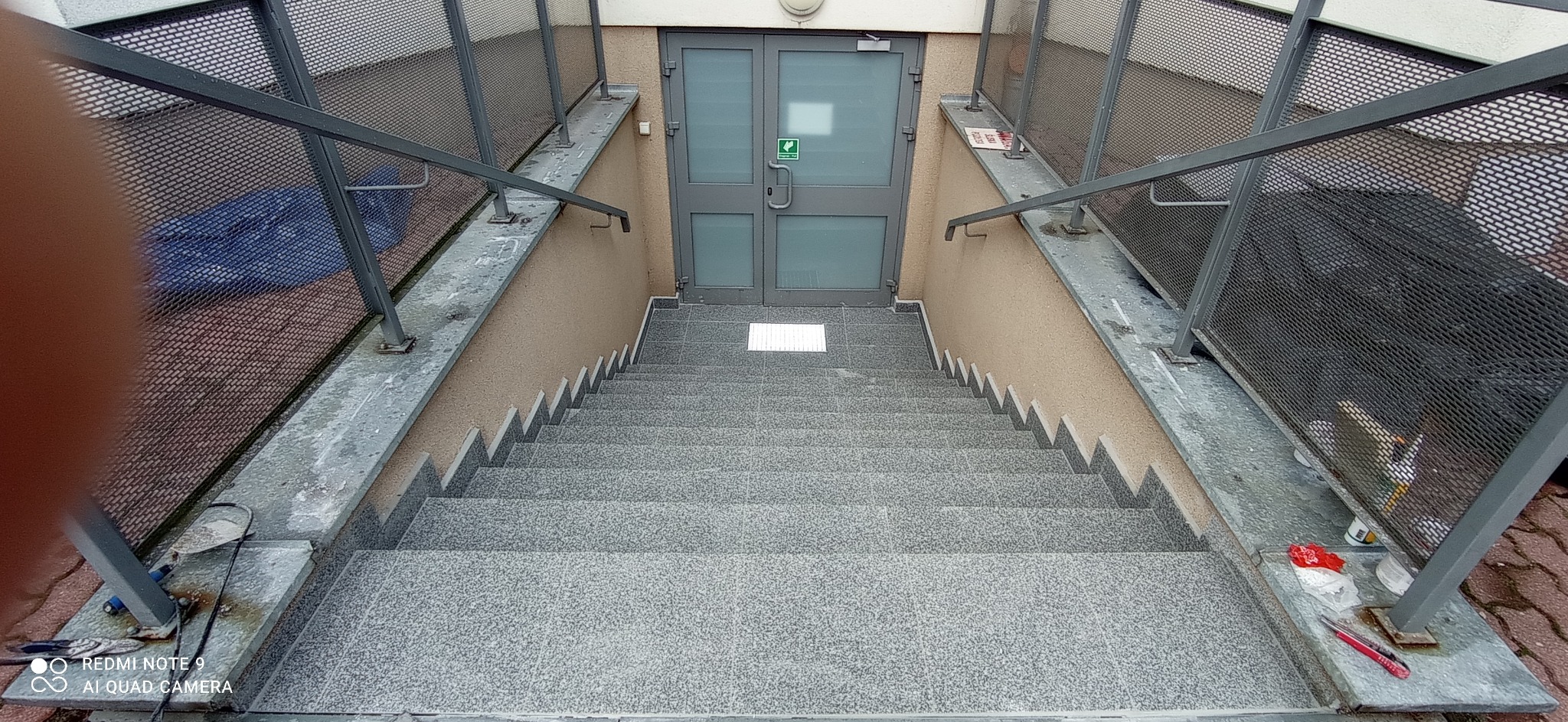 Zewnętrzne schody z szarej kostki prowadzące do budynku z metalową balustradą, widoczne narzędzia i resztki materiałów po remoncie.