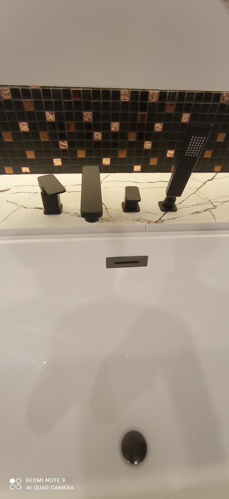 Czarna armatura łazienkowa z deszczownicą na tle mozaiki w odcieniach czerni i miedzi, zamontowana na marmurowym blacie w białej wannie.