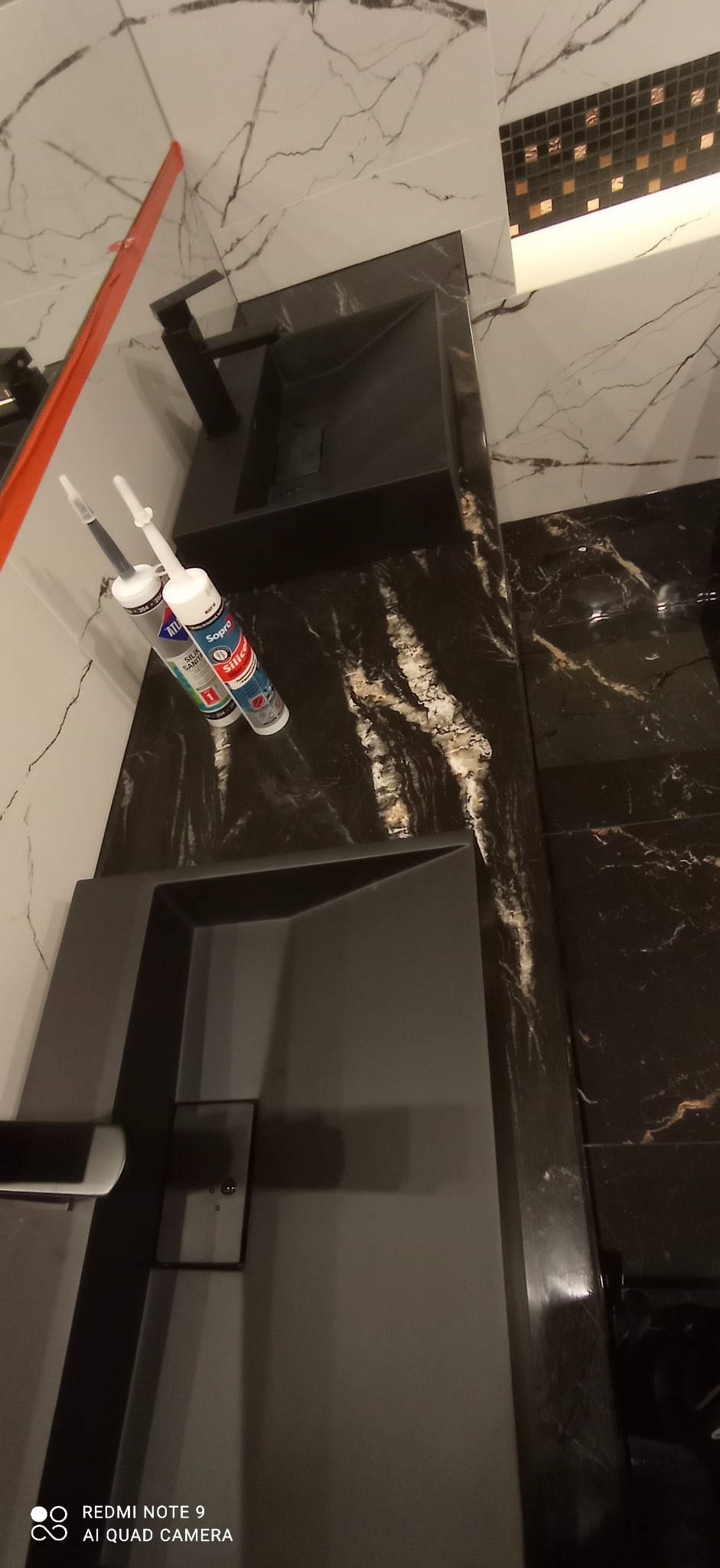 Czarny, nowoczesny zlew w łazience z marmurowymi płytkami na ścianach i podłodze, widoczne tuby silikonu Atlas i Sopro, czerwona poziomica przy ścianie.