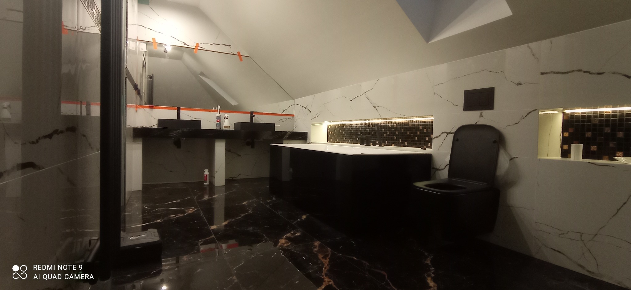 Nowoczesna łazienka na poddaszu z czarnymi elementami: wanna, umywalki, toaleta. Białe płytki z marmurowym wzorem na ścianach i podłodze. Dekoracyjna mozaika w niszy nad wanną.