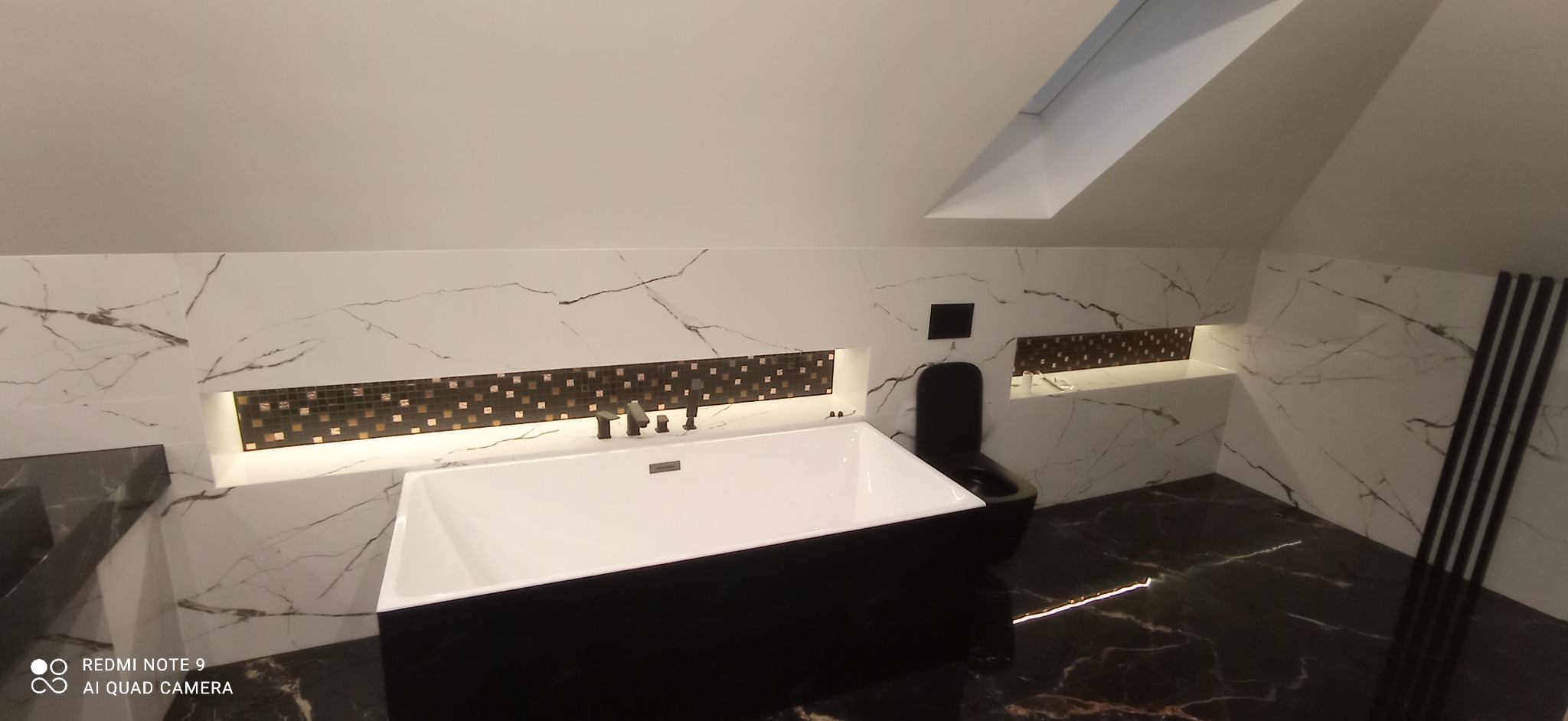 Nowoczesna łazienka na poddaszu z czarno-białymi marmurowymi płytkami, czarną wanną wolnostojącą i toaletą w tym samym kolorze, akcentowana mozaiką i oświetleniem LED w niszach.