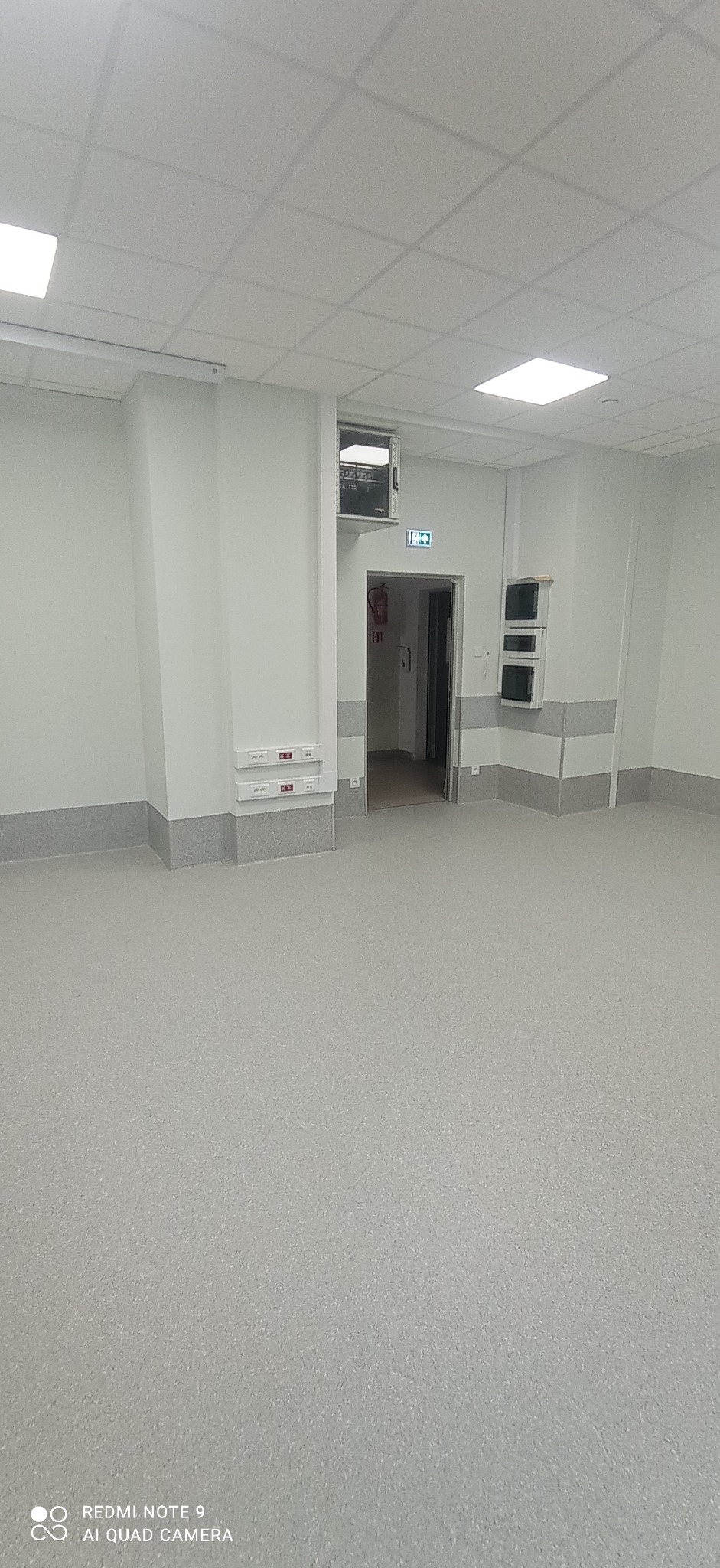 Wnętrze biurowe po remoncie: szara podłoga, białe ściany z szarym cokołem, sufit kasetonowy, widoczne gniazdka elektryczne i otwarte drzwi ewakuacyjne z gaśnicą.