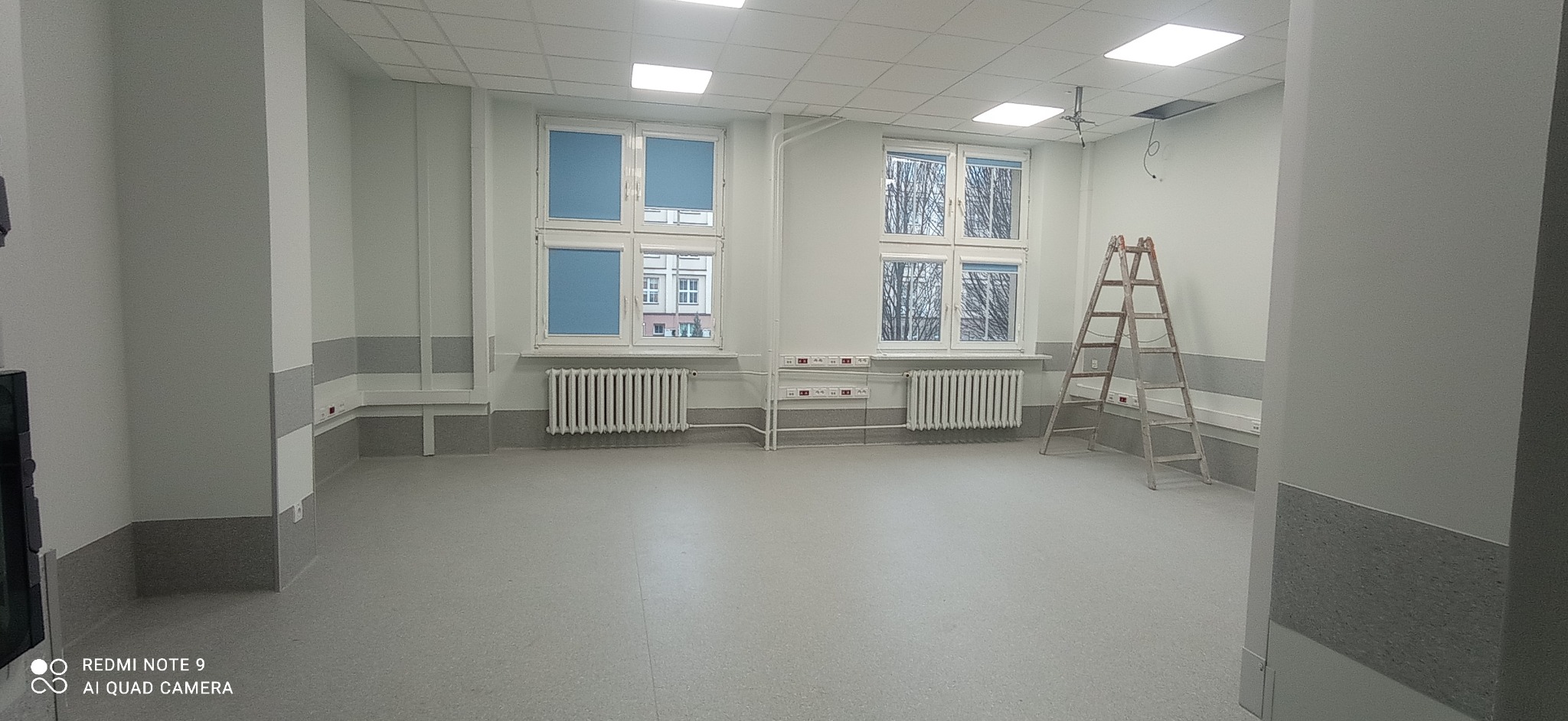 Pomalowane na jasno pomieszczenie biurowe z dwoma oknami z niebieskimi roletami, grzejnikami, instalacją elektryczną i aluminiową drabinką.