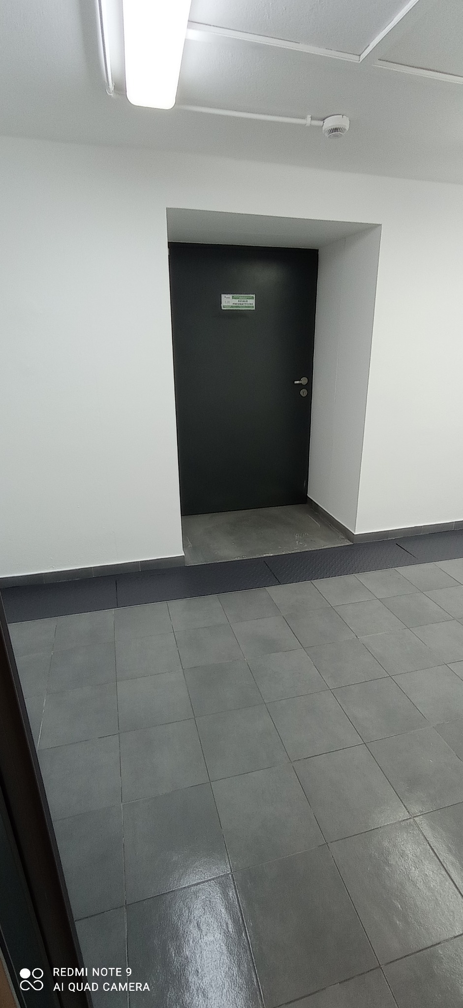 Szare płytki podłogowe w korytarzu biurowym, widoczna czarna ściana z drzwiami i tabliczką informacyjną.