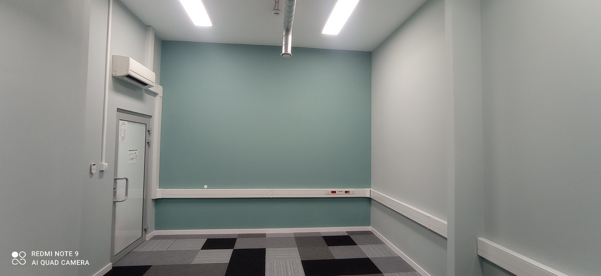 Wnętrze biurowe po malowaniu ścian na dwa odcienie szarości i zieleni, widoczna klimatyzacja, listwy przypodłogowe i sufitowe oraz wykładzina w szaro-czarną kratę.
