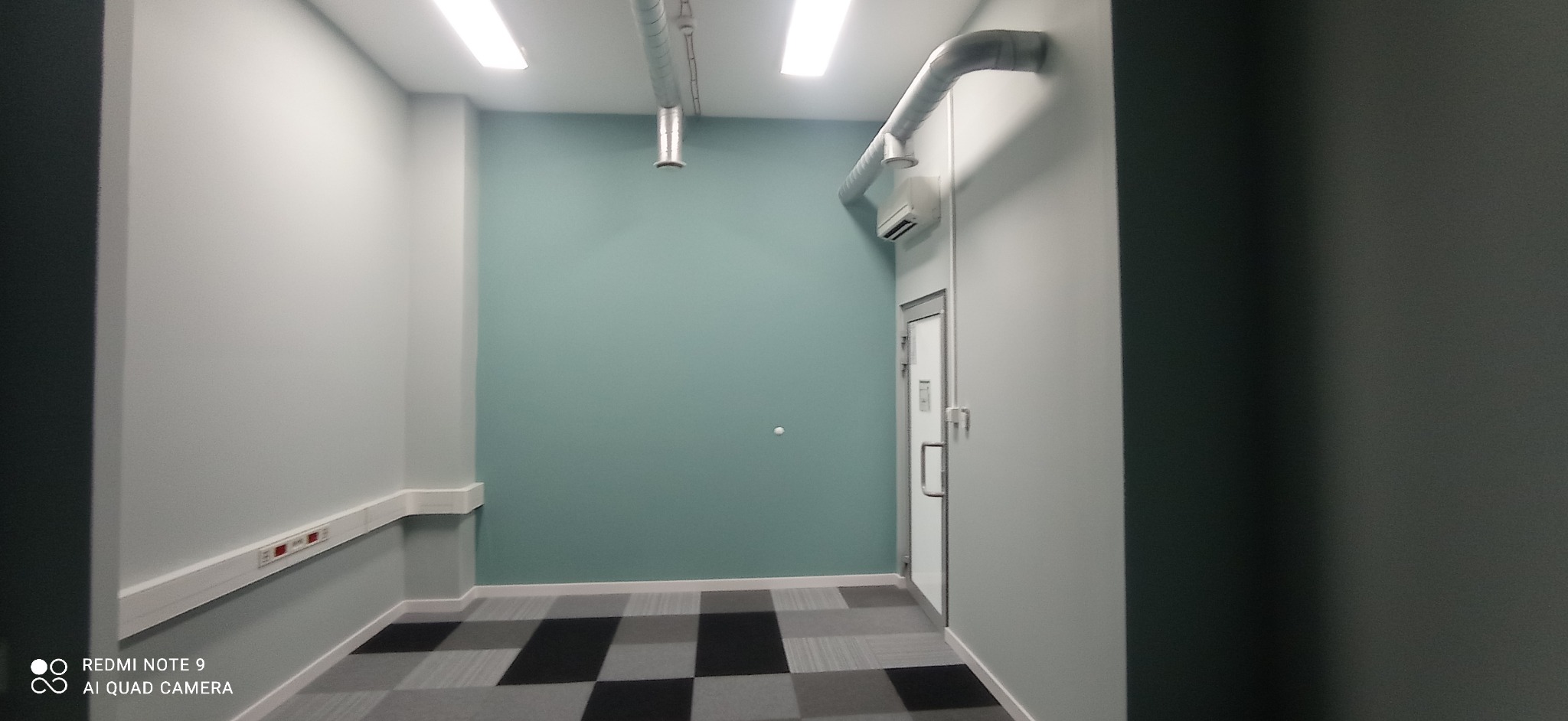 Wnętrze biura z pomalowanymi ścianami na szaro i turkusowo, widoczne instalacje wentylacyjne pod sufitem, szara podłoga w kratę i białe listwy przypodłogowe.