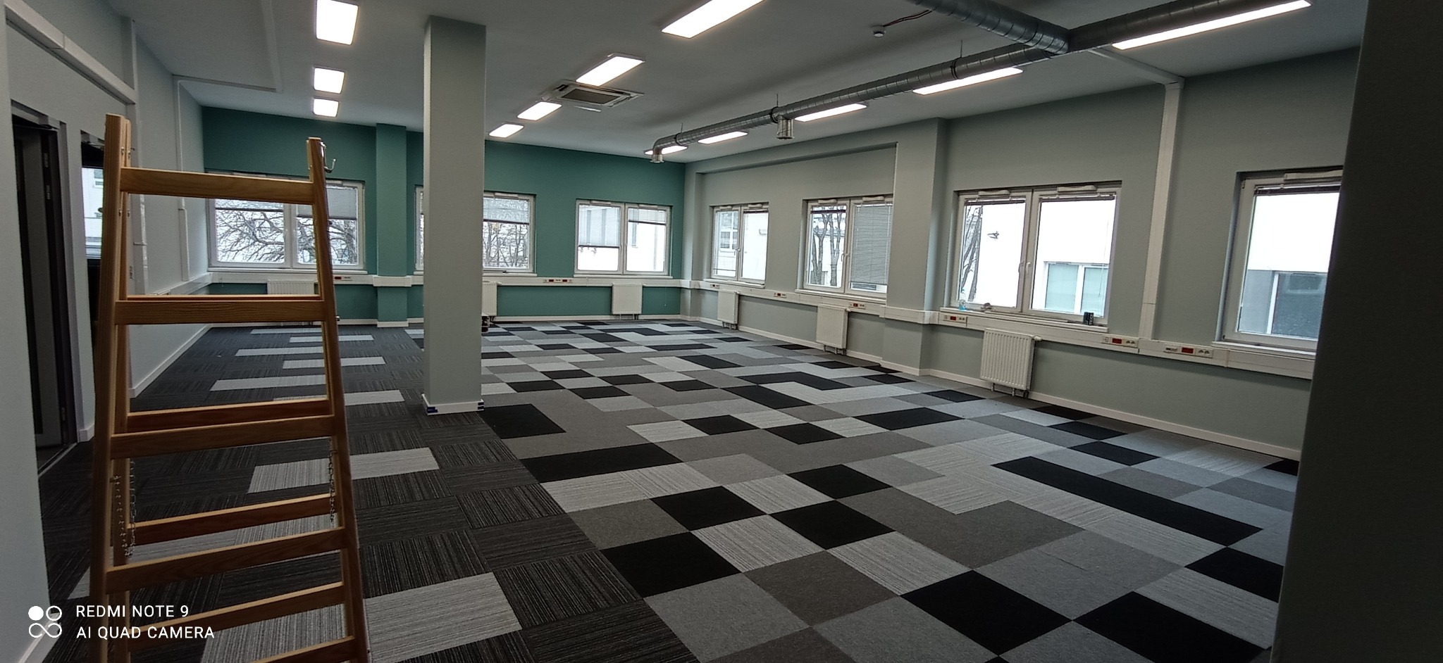 Przestronne biuro z nową, wzorzystą wykładziną dywanową w odcieniach szarości i czerni, drabina malarska w lewym rogu, ściany pomalowane na jasny odcień zieleni, widoczne grzejniki pod oknami.