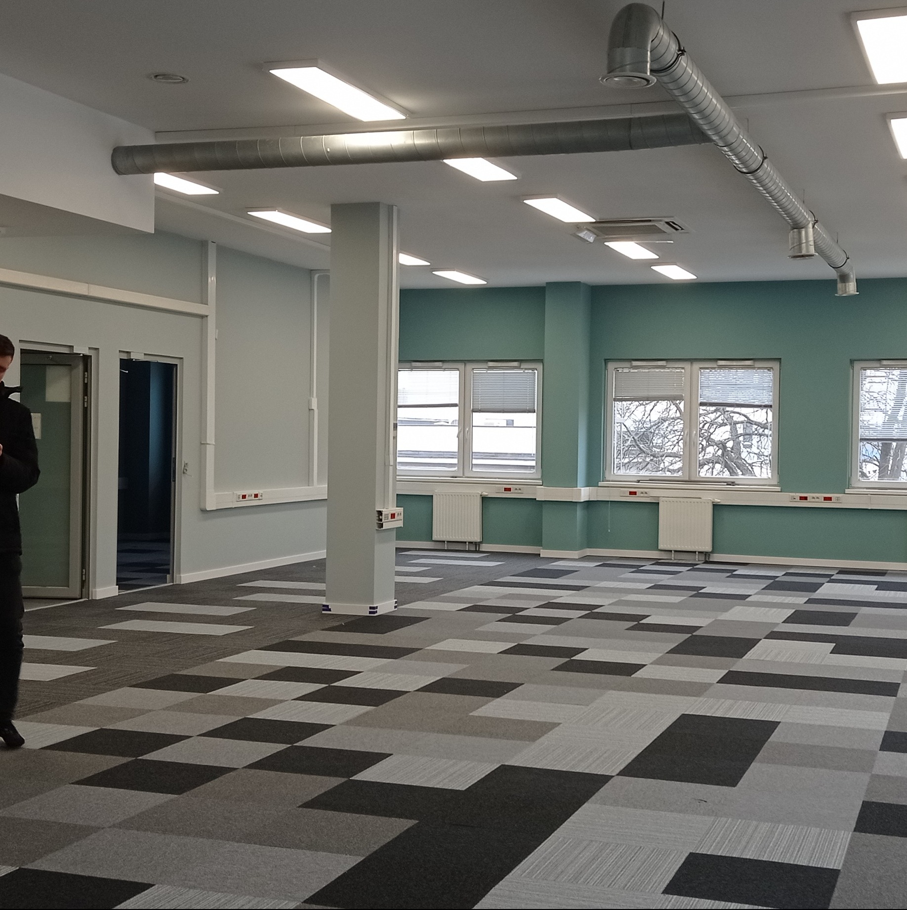 Pusta przestrzeń biurowa z szarą podłogą w kratę, turkusowymi ścianami, grzejnikami pod oknami i widocznymi rurami wentylacyjnymi pod sufitem.