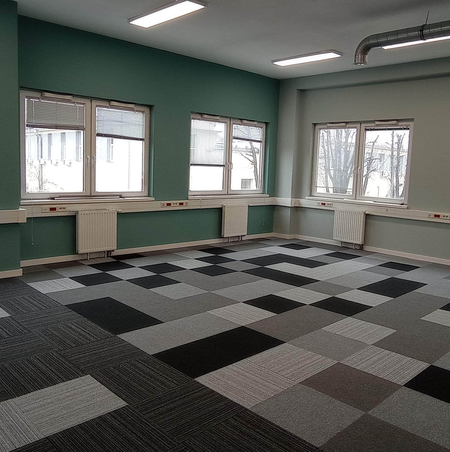 Pusta przestrzeń biurowa z panelami podłogowymi w odcieniach szarości i czerni, ściany pomalowane na zielono i szaro, trzy okna z żaluzjami, grzejniki pod oknami.