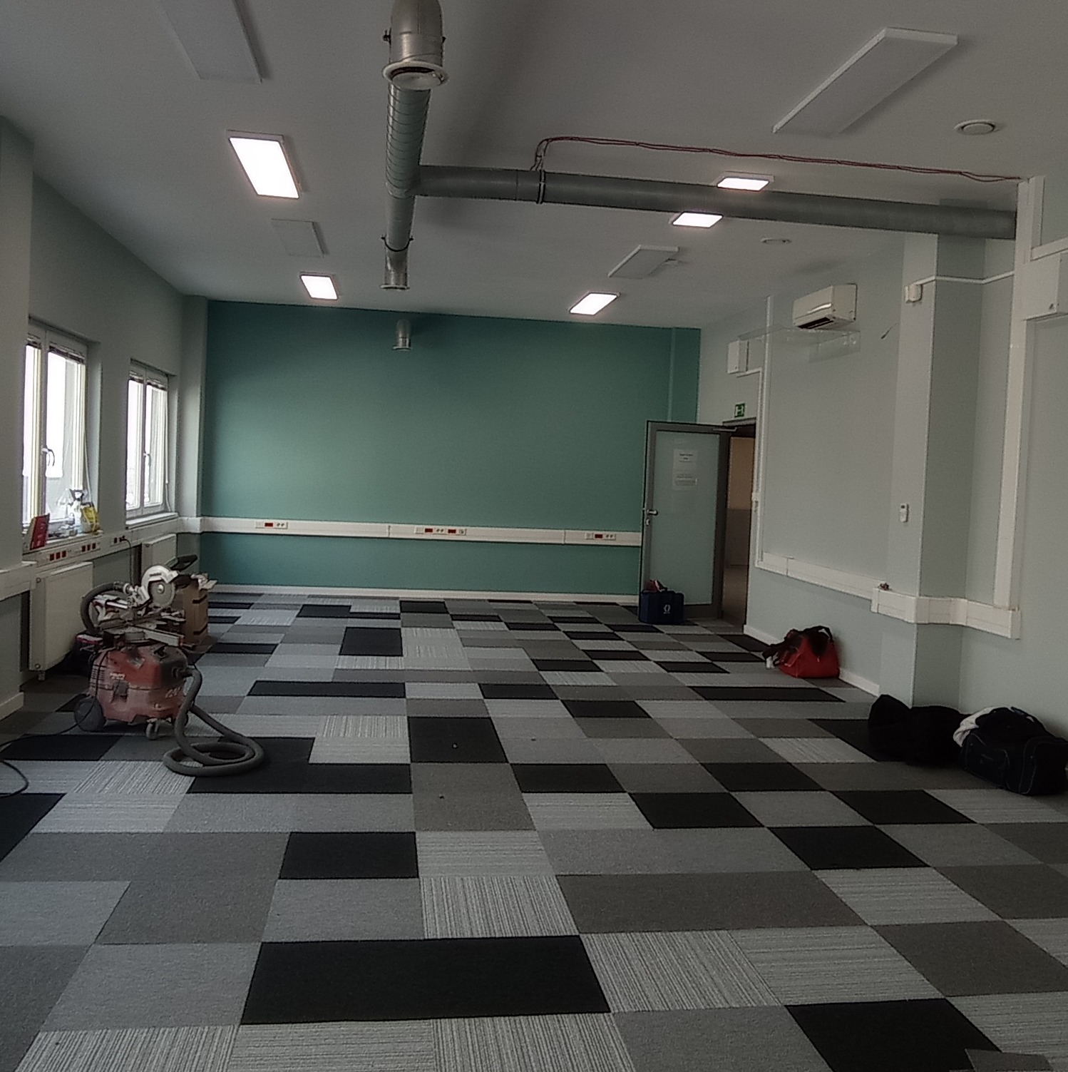 Wnętrze biura z szachownicową wykładziną dywanową w odcieniach szarości i czerni, przygotowane do malowania: zabezpieczone ściany, sprzęt malarski i odkurzacz przemysłowy w rogu pomieszczenia...