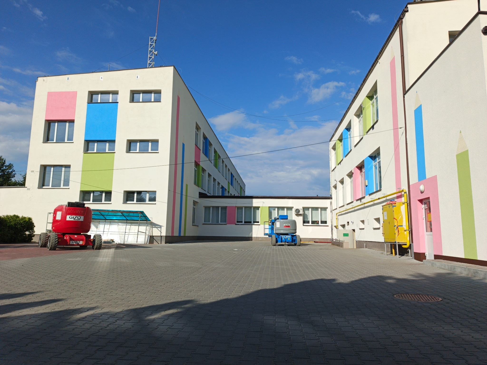 Budynek po malowaniu elewacji z kolorowymi akcentami i motywem kredek, widoczne podnośniki koszowe. Jasne, słoneczne ujęcie z szerokiej perspektywy.