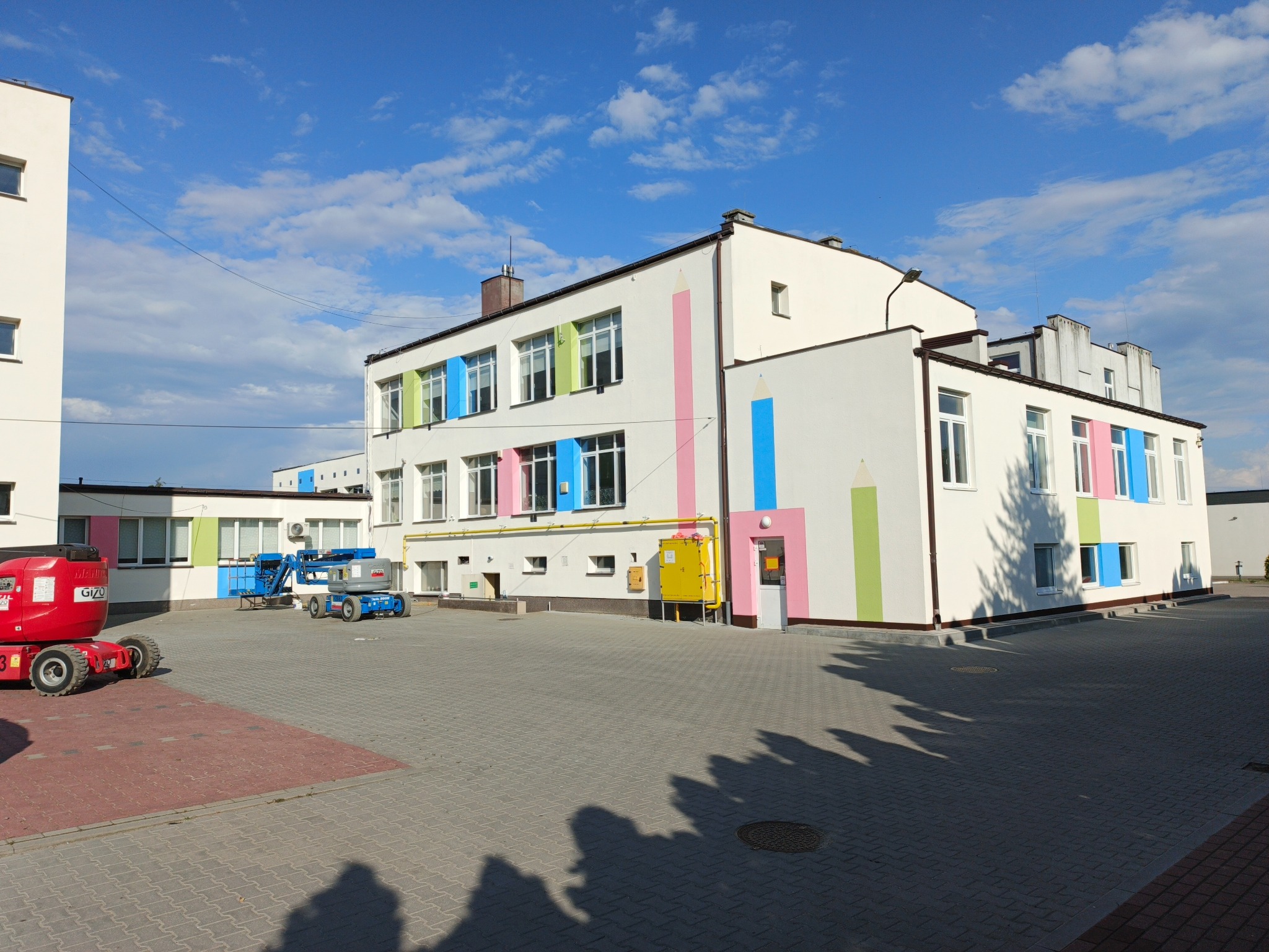 Biały budynek z kolorowymi oknami i rysunkami kredek, z podnośnikami koszowymi na dziedzińcu i błękitnym niebem w tle.