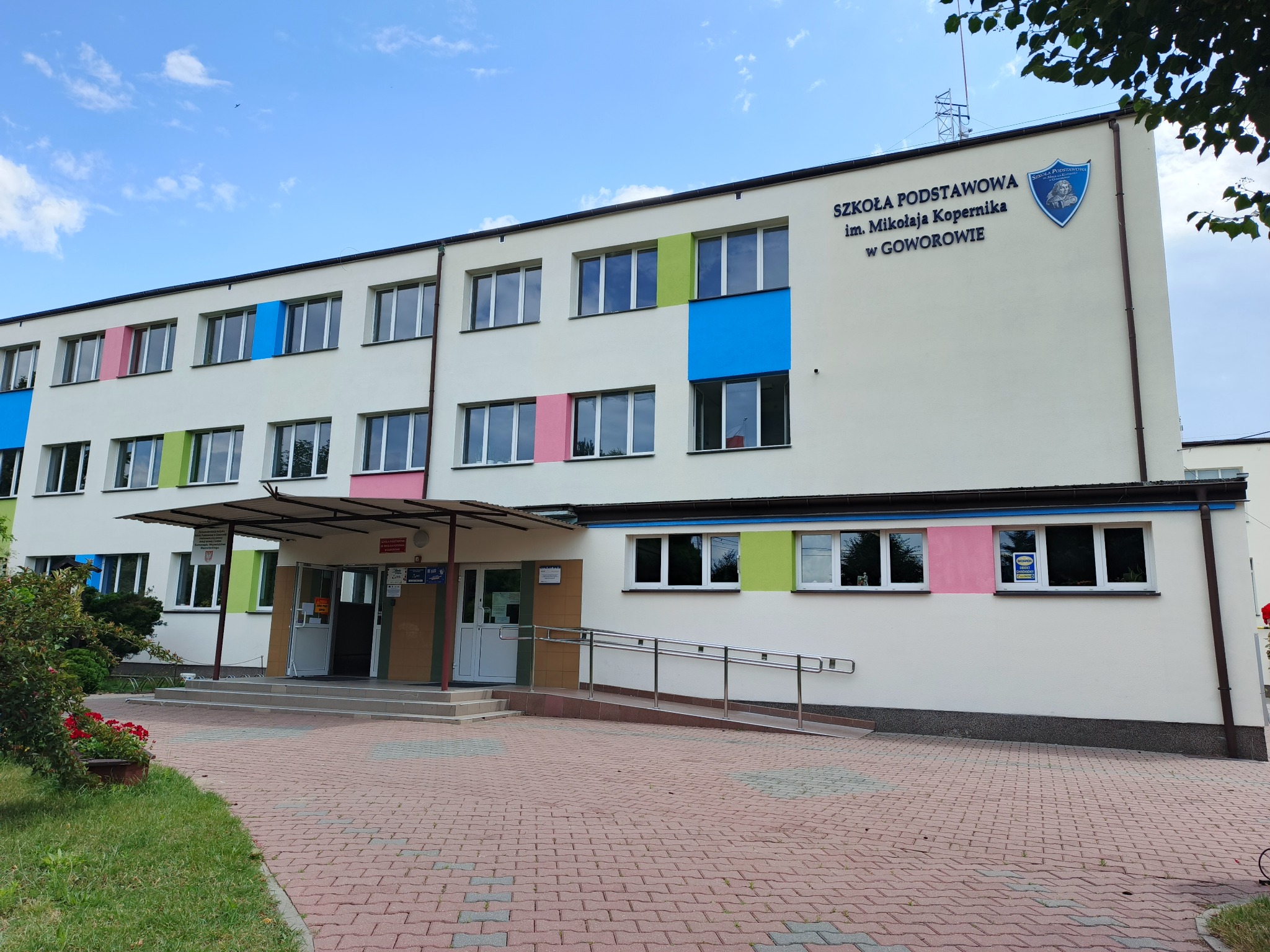 Fasada budynku szkoły podstawowej z kolorowymi akcentami wokół okien: niebieski, zielony, różowy. Wejście z podjazdem dla wózków. Czerwona kostka brukowa.