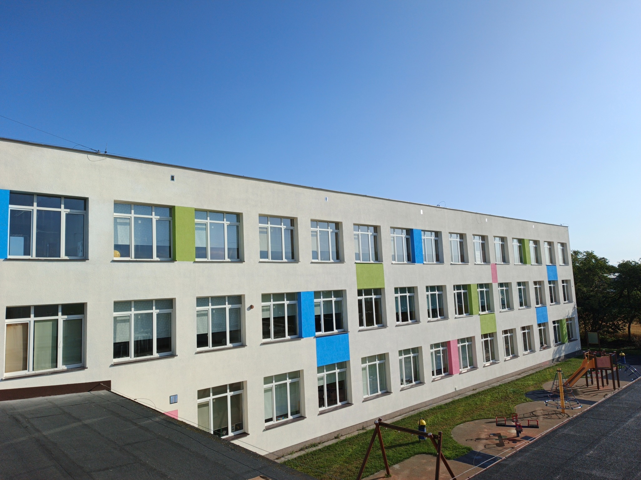 Fasada budynku z oknami, fragment dachu i plac zabaw w tle. Okna ozdobione kolorowymi panelami: niebieskim, zielonym i różowym. Jasne, słoneczne oświetlenie.
