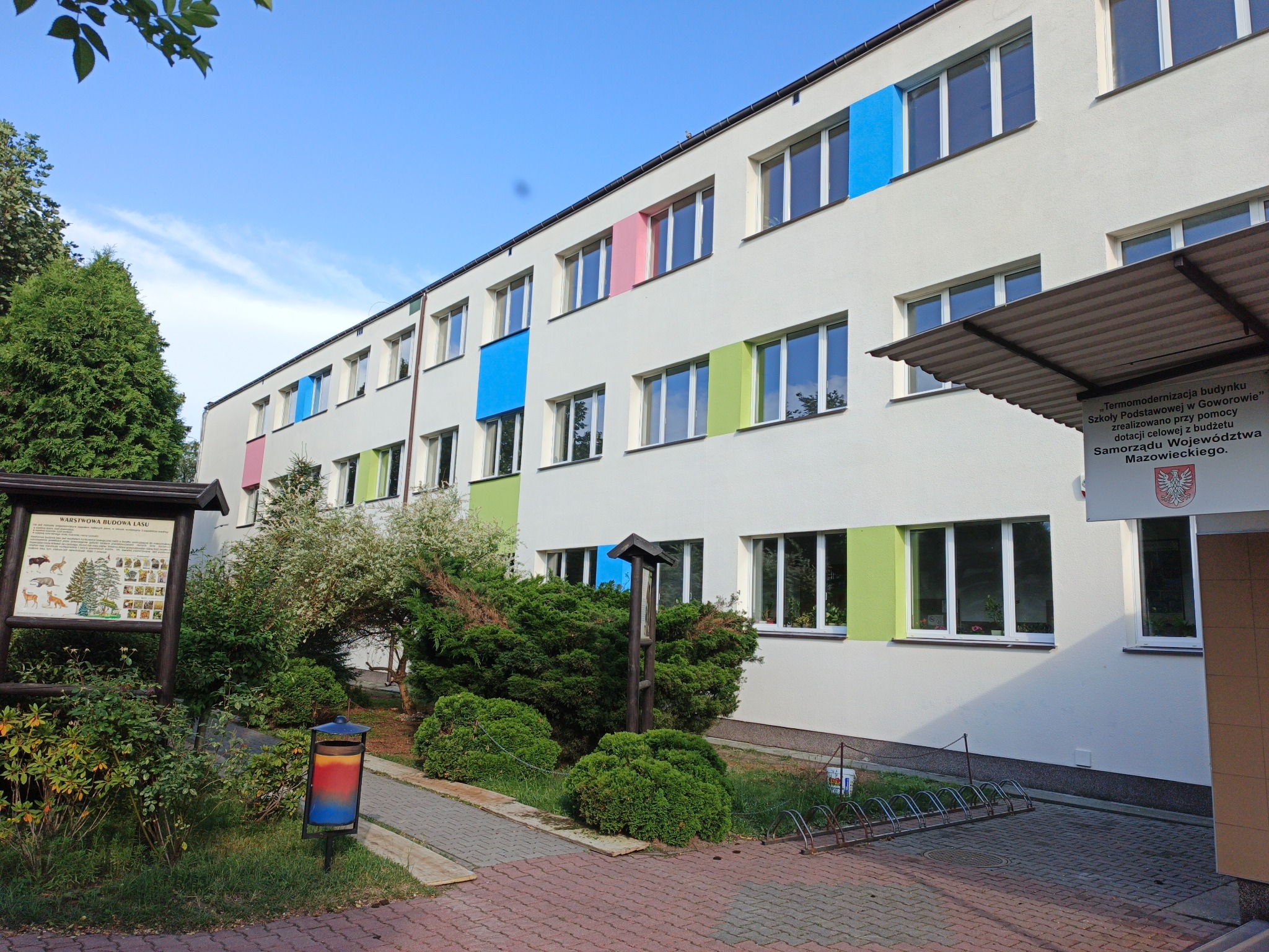 Budynek szkoły po termomodernizacji z kolorowymi obramowaniami okien: niebieskie, zielone, różowe. Widoczny fragment chodnika i zieleń. Tablica informacyjna.