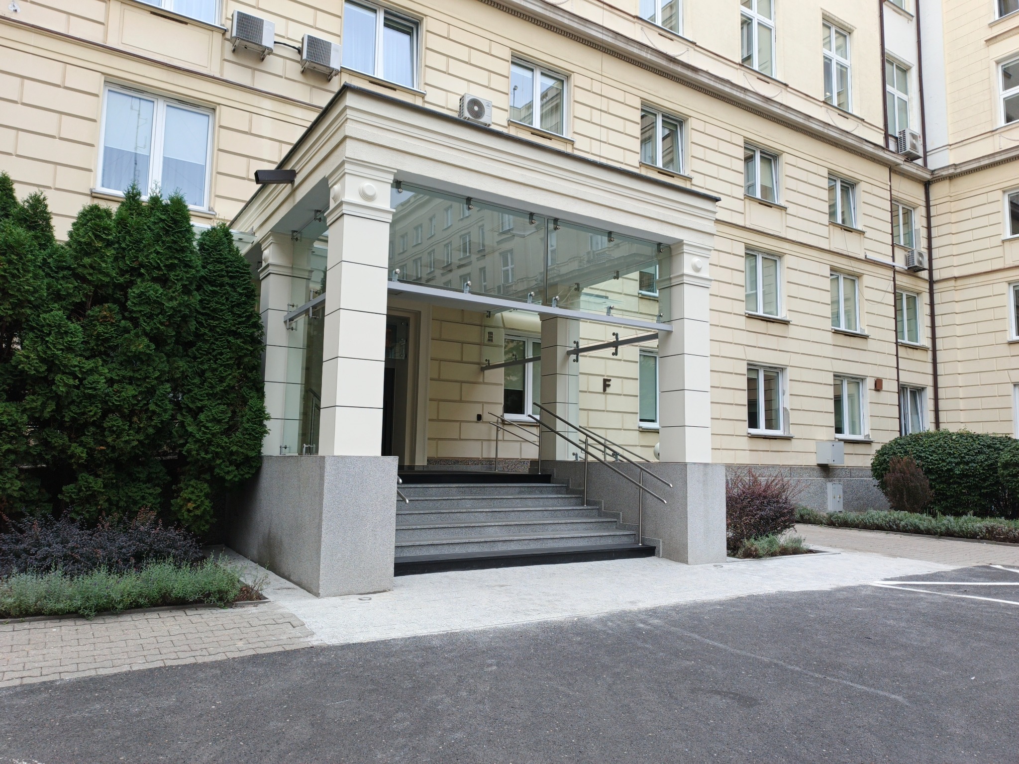 Wejście do budynku z przeszklonym zadaszeniem i granitowymi schodami. Elewacja w odcieniach beżu, widoczne okna, klimatyzatory. Obok wejścia zieleń.