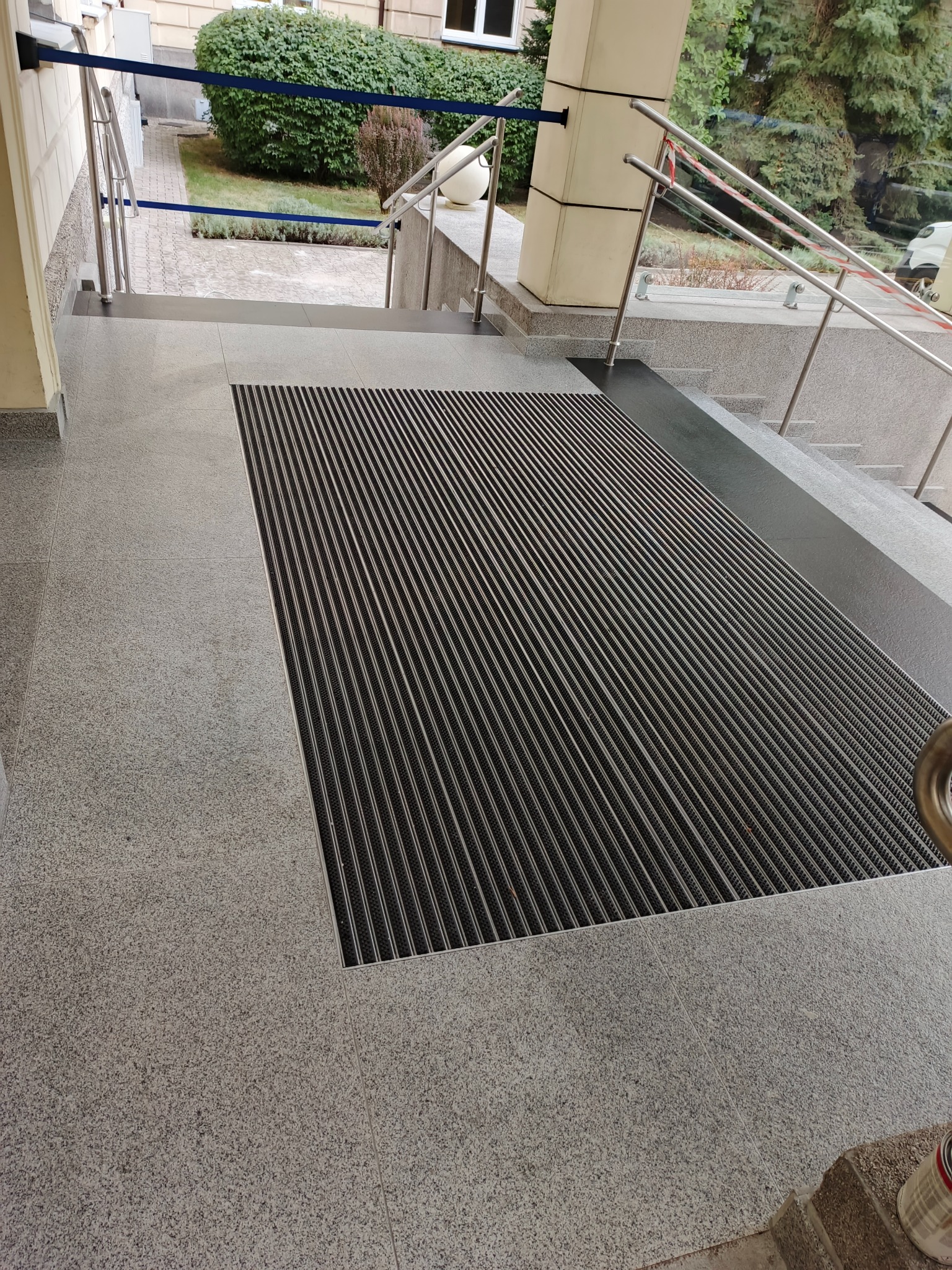 Szare płytki podłogowe z czarną wycieraczką systemową na wejściu do budynku. Widoczne schody z barierkami i roślinność w tle.