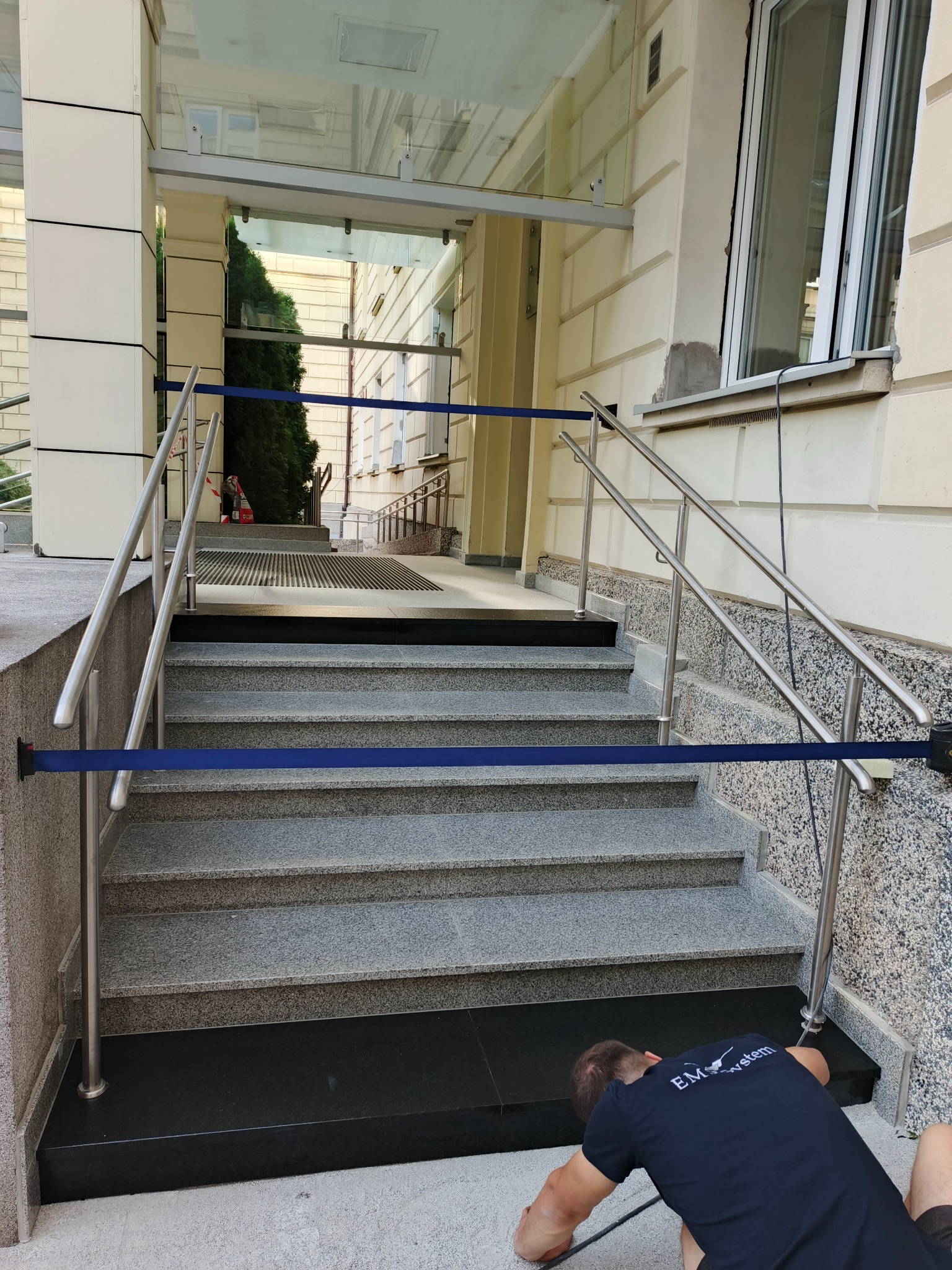 Granitowe schody wejściowe z barierkami zabezpieczone taśmą, w trakcie montażu przez pracownika w koszulce z logo firmy. Wykończenie elewacji budynku w tle.