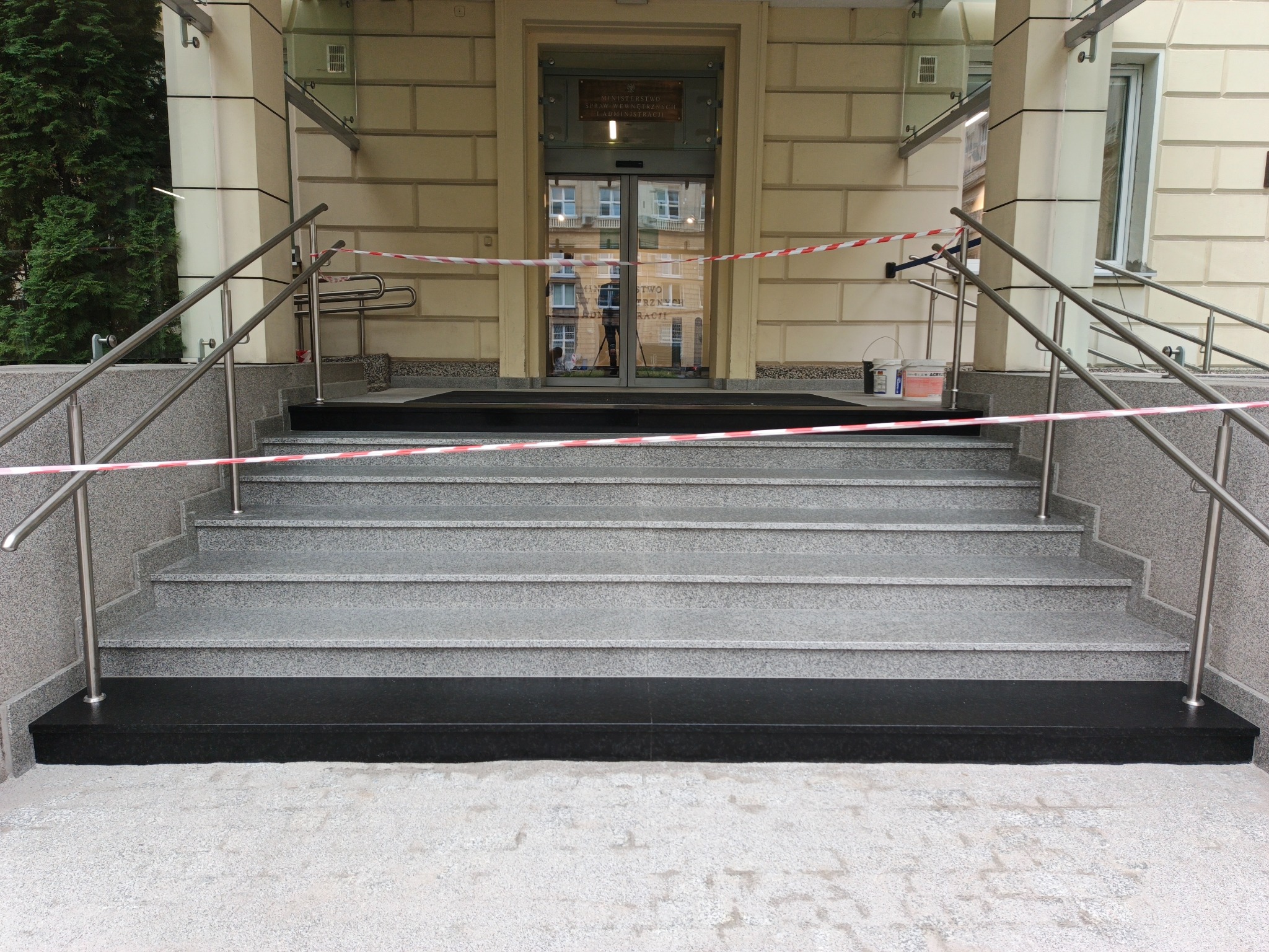 Granitowe schody wejściowe do budynku z barierkami, zabezpieczone taśmą ostrzegawczą. Widoczne ślady po pracach wykończeniowych i wiadro z farbą.