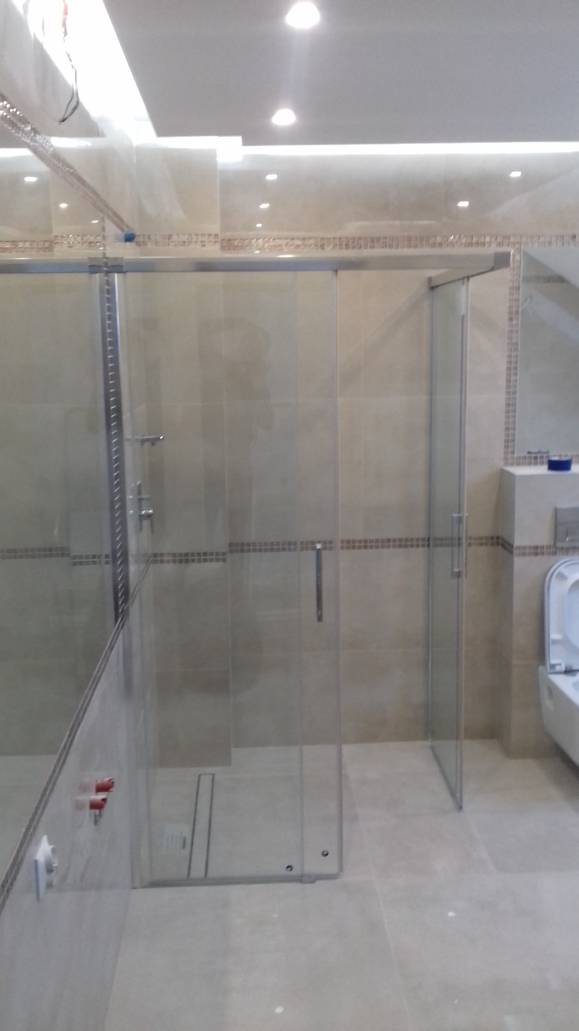 Szklana kabina prysznicowa z przesuwnymi drzwiami w beżowej łazience z mozaikowym pasem, widoczny fragment toalety.