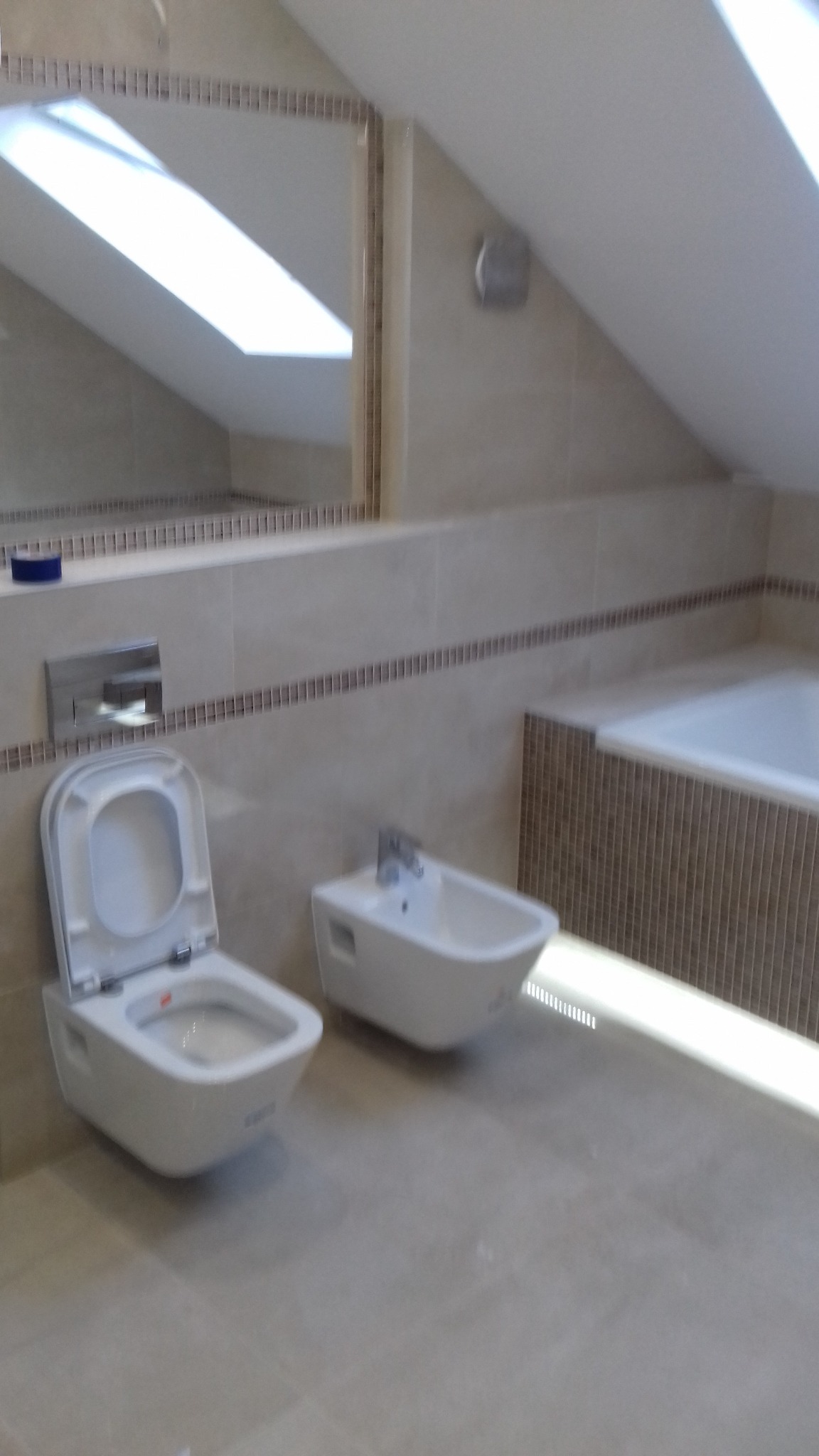 Nowoczesna łazienka na poddaszu z beżowymi płytkami, wiszącą toaletą z otwartą klapą, bidetem i lustrem odbijającym okno dachowe.