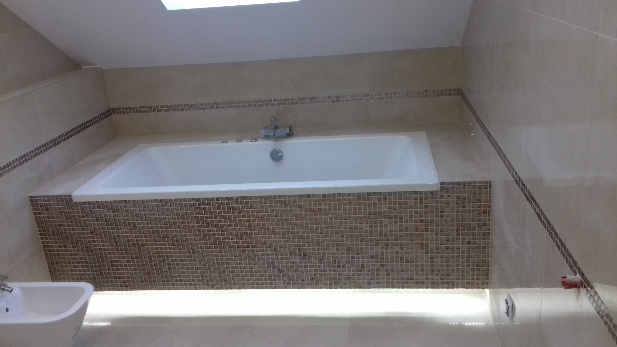 Wykończona łazienka na poddaszu: biała wanna obudowana mozaiką, kremowe płytki na ścianach z dekoracyjnym paskiem mozaiki, bidet w rogu.