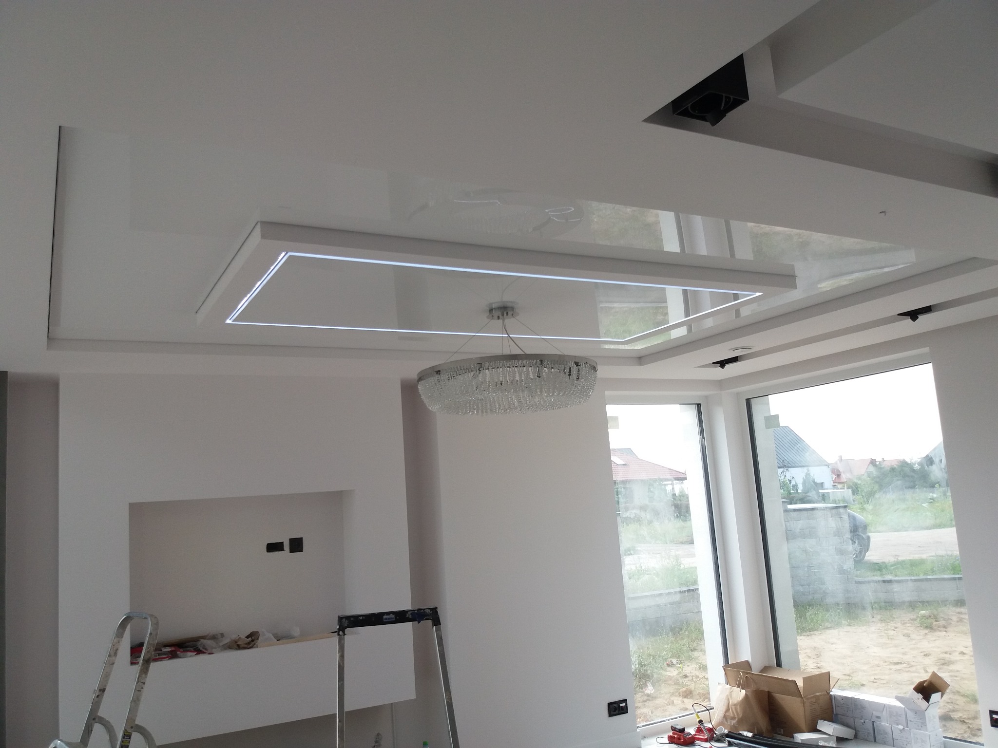 Nowoczesny sufit podwieszany z oświetleniem LED w kształcie prostokąta i kryształowym żyrandolem, widoczne drabiny i kartony sugerujące prace wykończeniowe w jasnym wnętrzu.