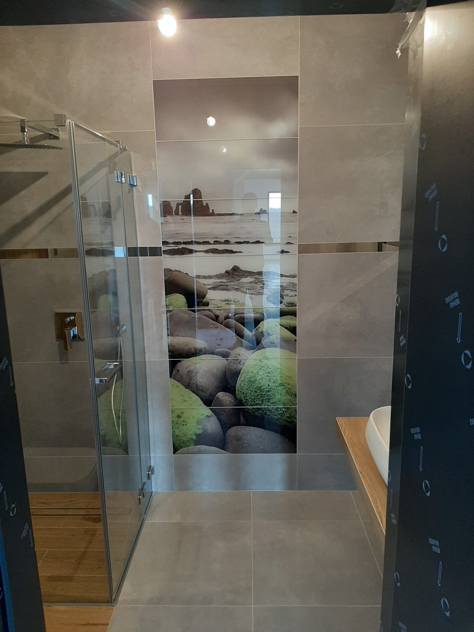 Szare płytki w łazience z motywem kamieni i morza, kabina prysznicowa ze złotymi elementami, drewniany blat pod umywalką.