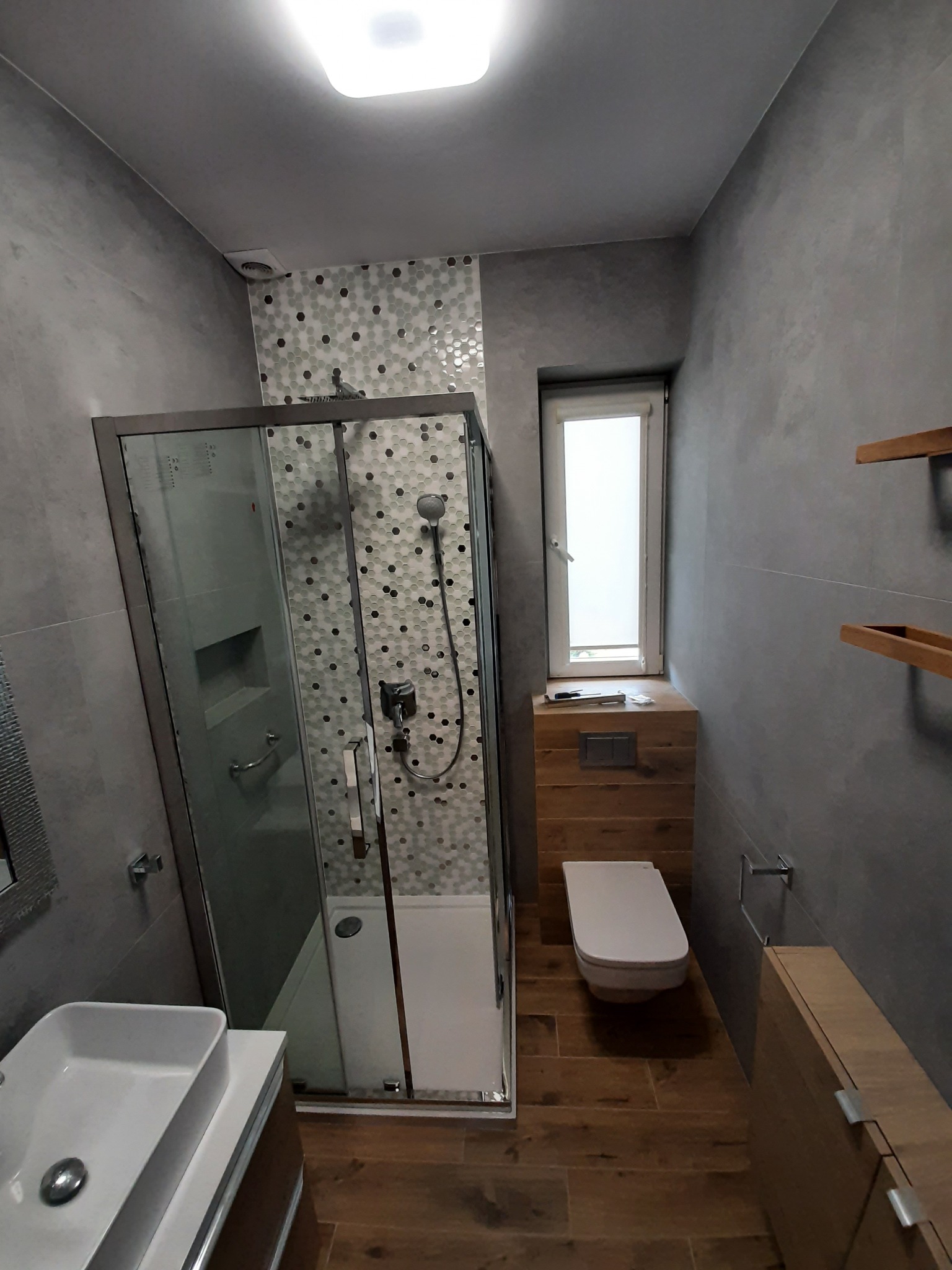 Nowoczesna łazienka z szarą, teksturowaną ścianą, kabiną prysznicową z mozaiką, wiszącą toaletą z drewnianą obudową i drewnianą podłogą.