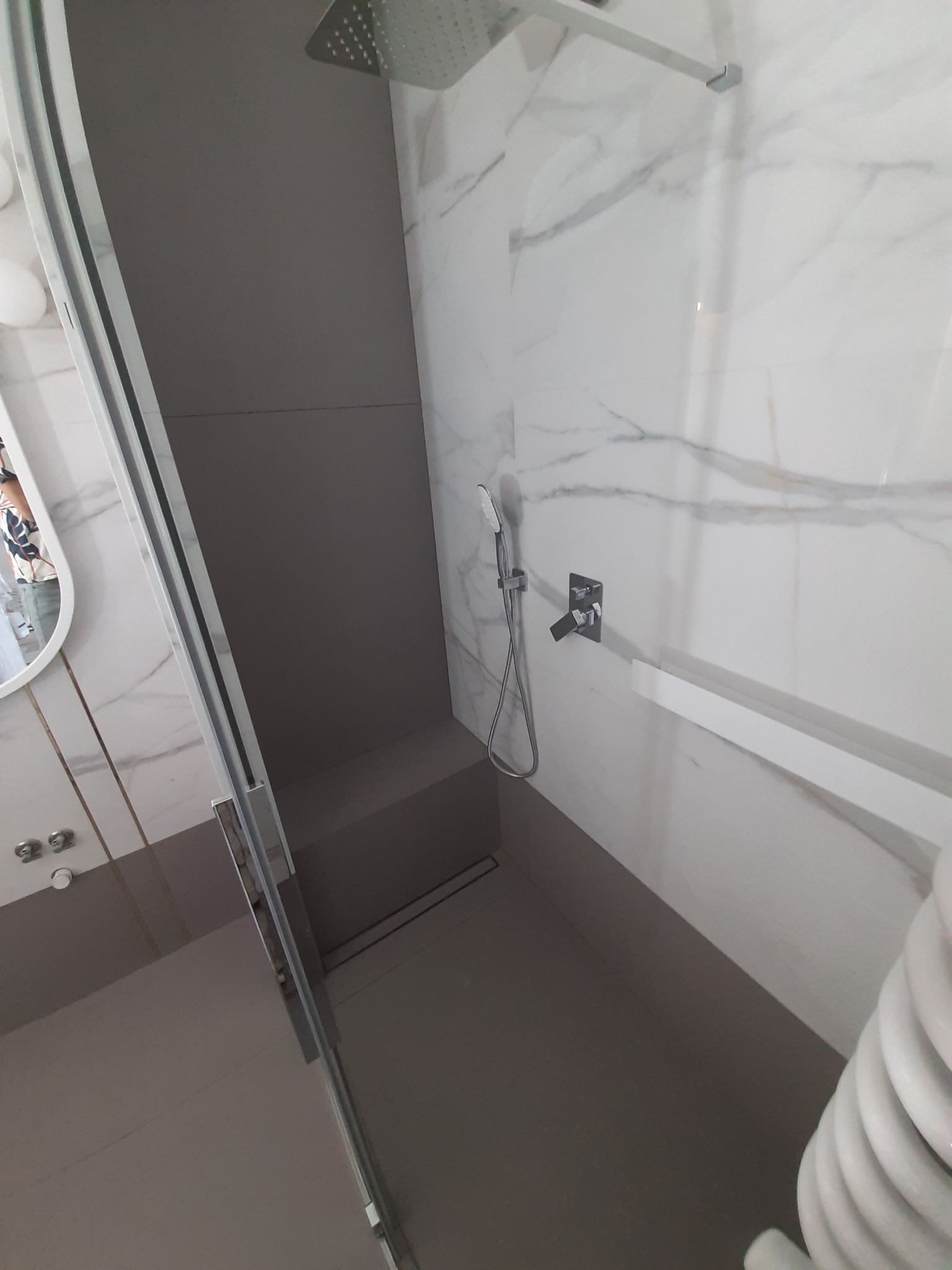 Nowoczesna kabina prysznicowa z deszczownicą i marmurowymi płytkami ściennymi, szara podłoga i ścianka boczna, widoczny fragment grzejnika.